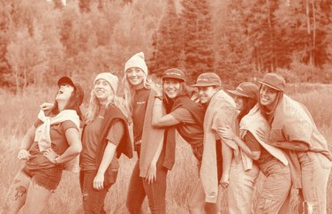 Group of friends in Fly Like A Girl caps and shirts laughing in a meadow.